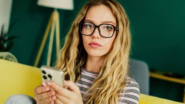 Young woman with long blonde hair and glasses sitting on a yellow couch, holding a smartphone, looking toward the camera.