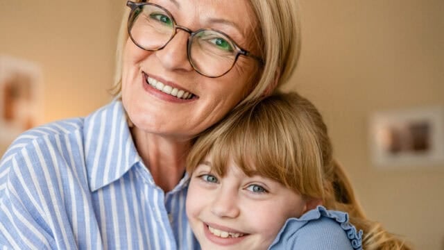 An older woman wearing glasses smiles while hugging a young girl with blonde hair. Both are happy and looking at the camera in a warmly lit room.