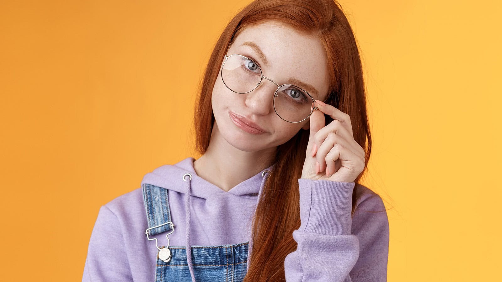 Young woman with long red hair, wearing glasses, a lavender hoodie, and denim overalls, looking at the camera with a slight smile and adjusting her glasses, standing against a bright orange background.