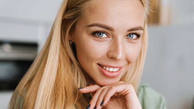 A woman with long blonde hair and blue eyes smiles gently at the camera, resting her chin on her hand.