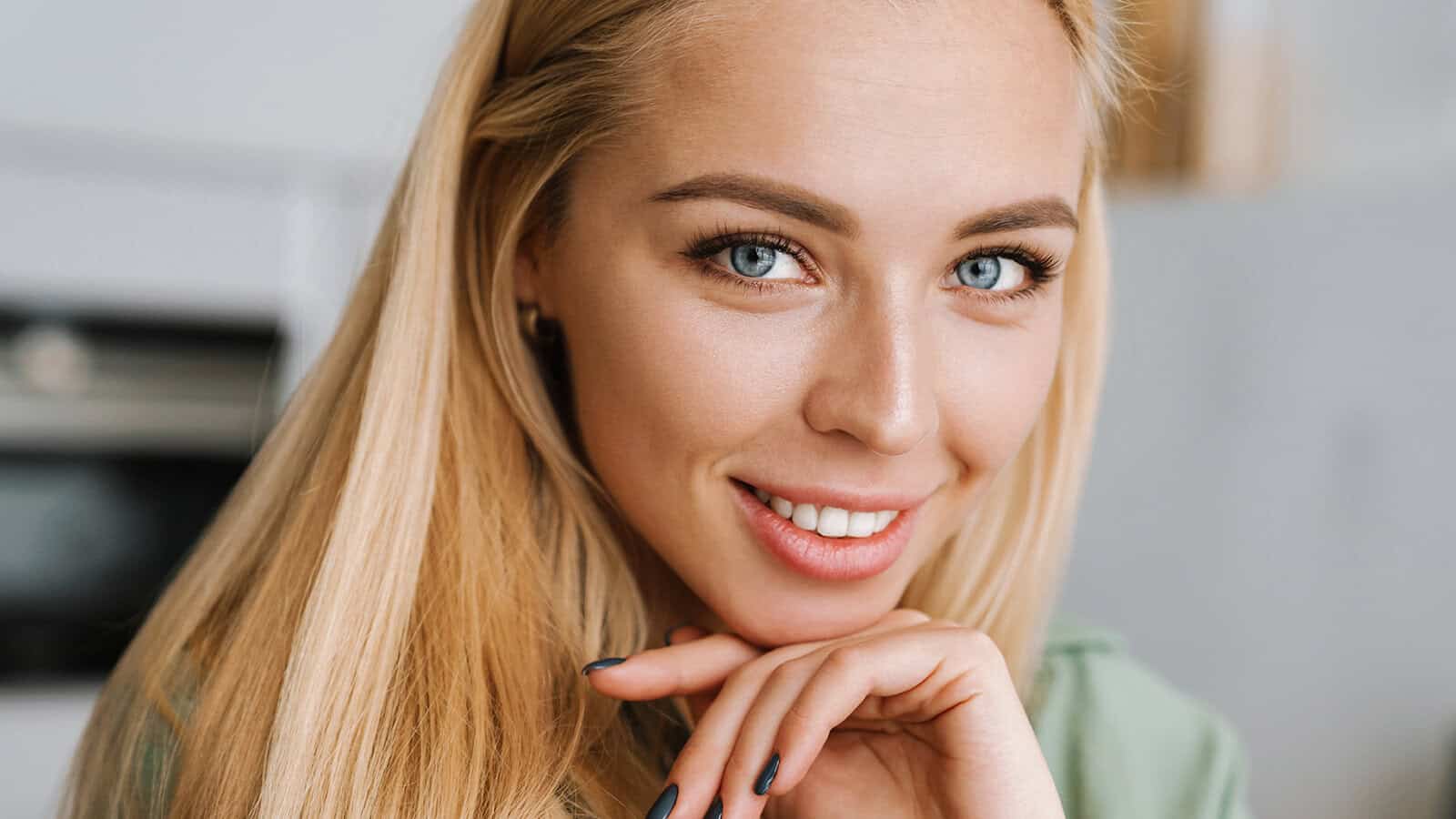 A woman with long blonde hair and blue eyes smiles gently at the camera, resting her chin on her hand.