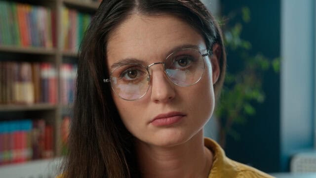 A woman with long brown hair and glasses looks directly at the camera with a neutral expression. She is indoors, with bookshelves and a plant in the blurred background.