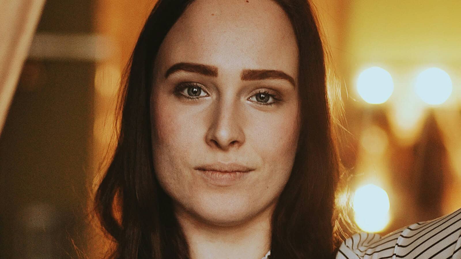 A person with long brown hair and strong eyebrows looks directly at the camera with a neutral expression; warm lighting and blurred lights are in the background.