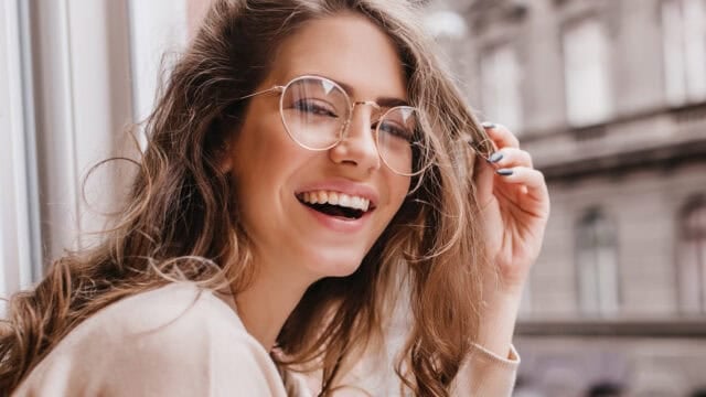 Smiling woman with long wavy hair and round glasses sits by a window, wearing a light sweater. She is looking at the camera and holding her hair with one hand. Blurred city buildings are visible outside.