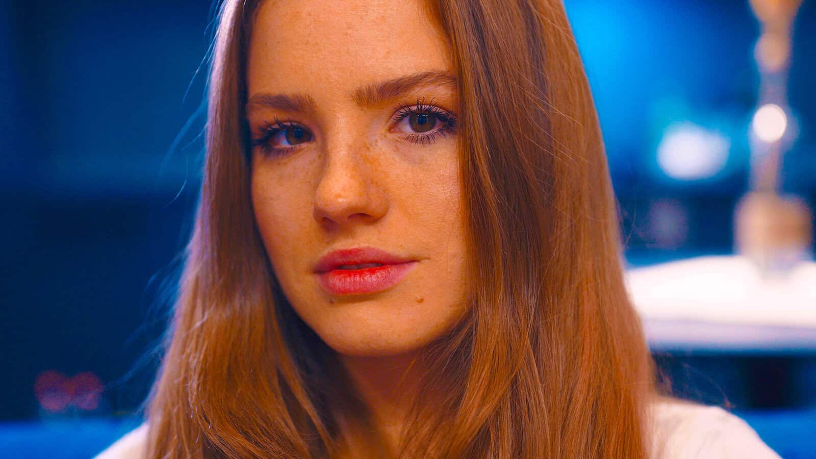 A young woman with long auburn hair and freckles looks directly at the camera with a neutral expression, set against a softly blurred blue background.