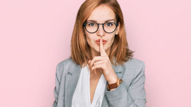 A woman with glasses and a gray blazer holds a finger to her lips, signaling shh or silence, against a light pink background.