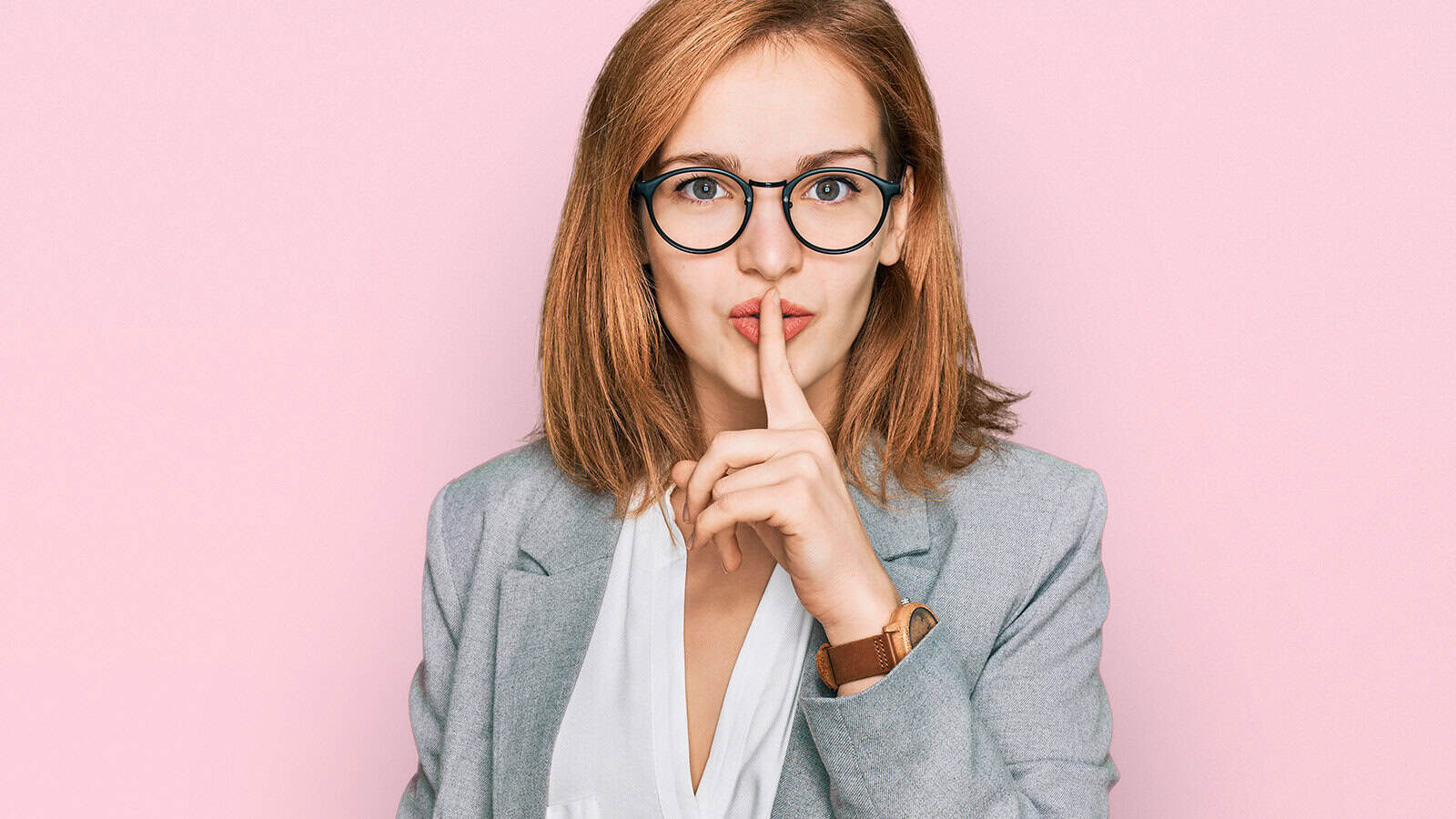 A woman with glasses and a gray blazer holds a finger to her lips, signaling shh or silence, against a light pink background.
