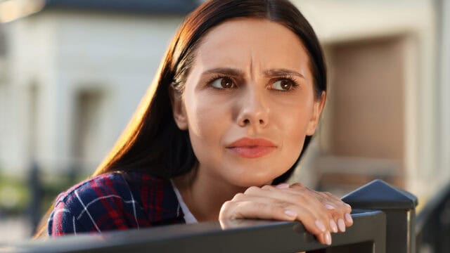 A woman with dark hair looks to the side with a concerned expression, leaning her hands on a metal fence outdoors.