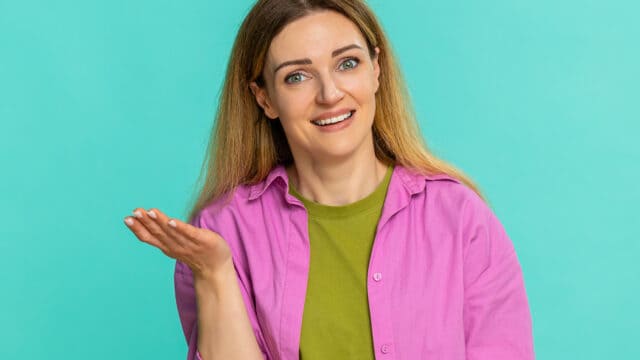 A woman with long, light brown hair smiles and gestures with one hand raised. She wears a green shirt and an open pink button-up shirt, standing against a plain turquoise background.