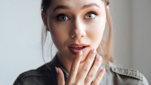 A young woman with light eyes and brown hair looks surprised, holding her hand to her mouth. She wears a green shirt and has natural makeup, standing in soft natural light.