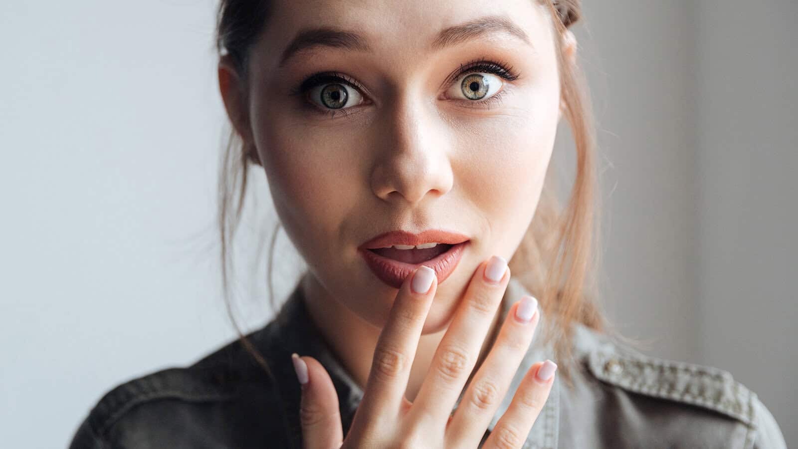 A young woman with light eyes and brown hair looks surprised, holding her hand to her mouth. She wears a green shirt and has natural makeup, standing in soft natural light.