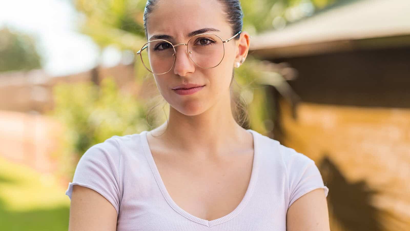 A young person with glasses and a white shirt stands outdoors with a serious expression, blurred greenery and a building in the background.