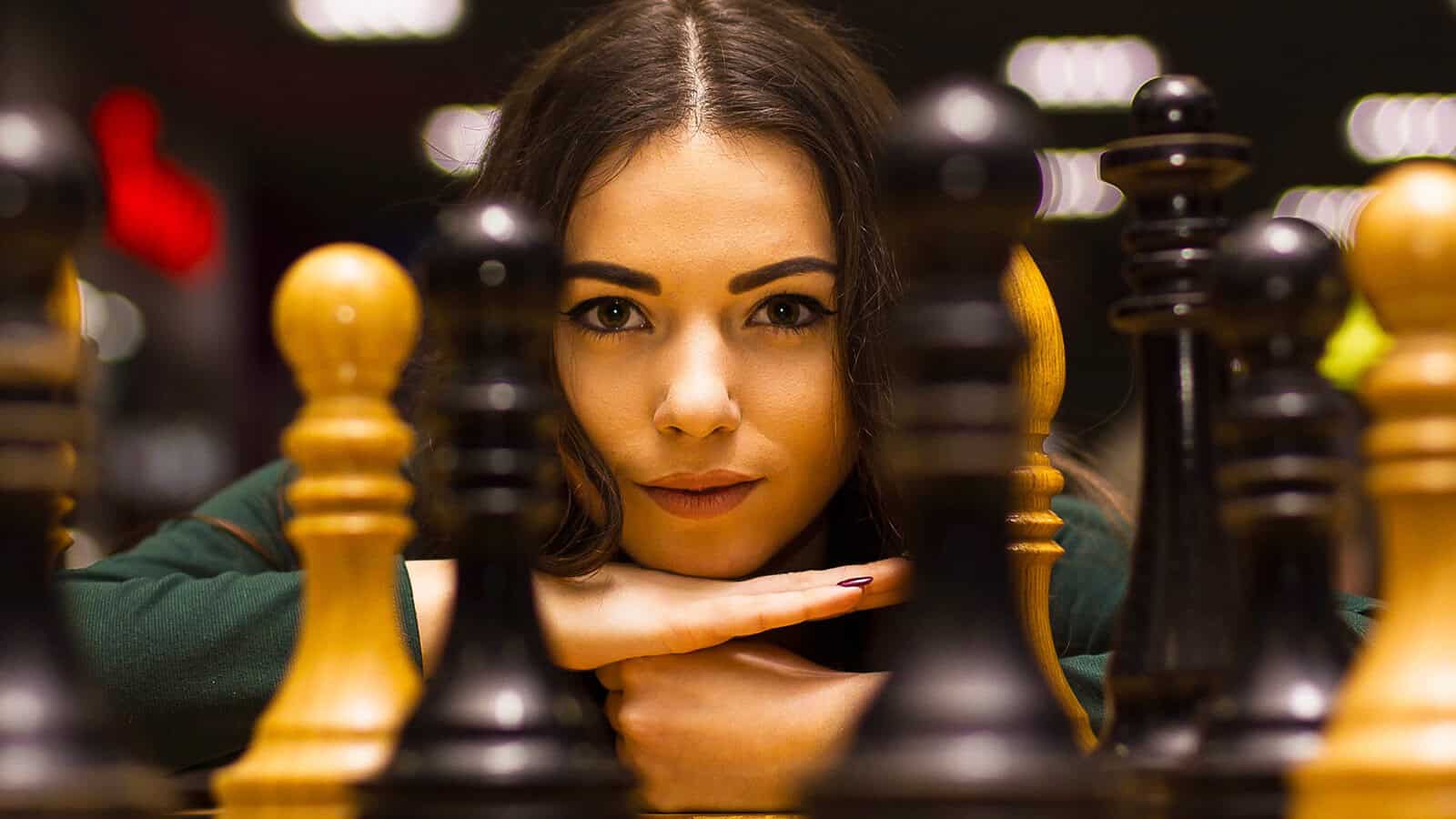 A woman with long brown hair rests her chin on her hands and looks forward, surrounded by black and white chess pieces on a chessboard.