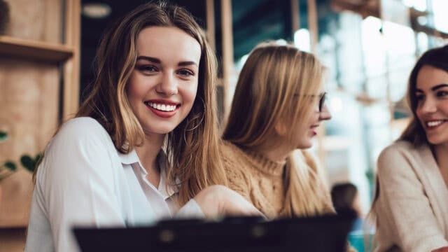 Three young women sit together in a cozy café. One woman in the foreground smiles brightly at the camera, while the others are engaged in conversation. Warm lighting and a relaxed atmosphere fill the scene.