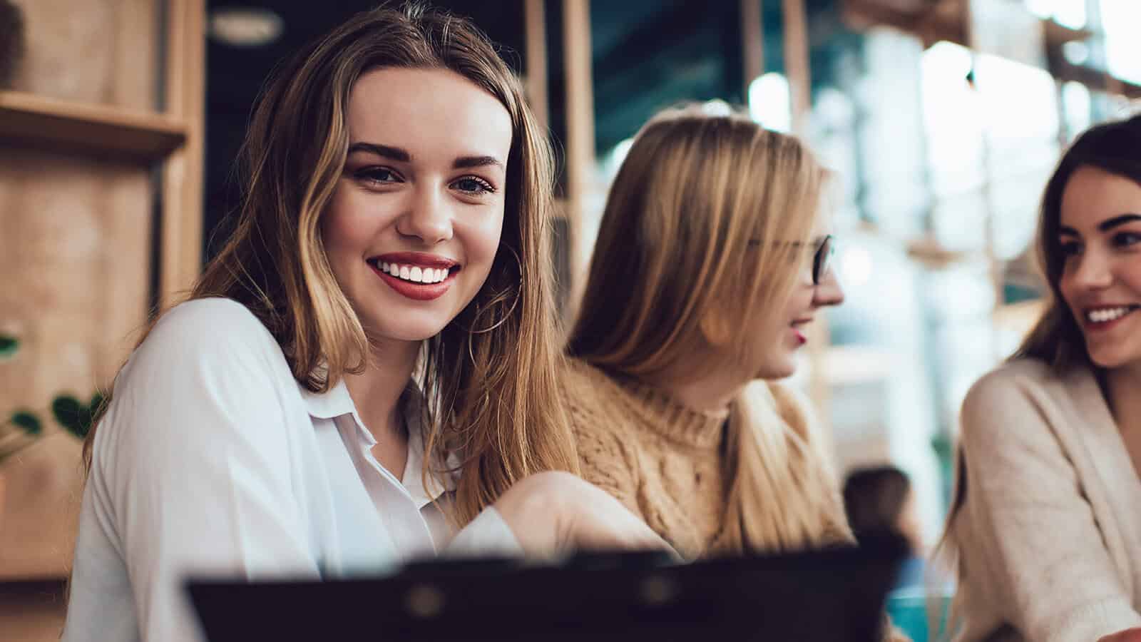 Three young women sit together in a cozy café. One woman in the foreground smiles brightly at the camera, while the others are engaged in conversation. Warm lighting and a relaxed atmosphere fill the scene.