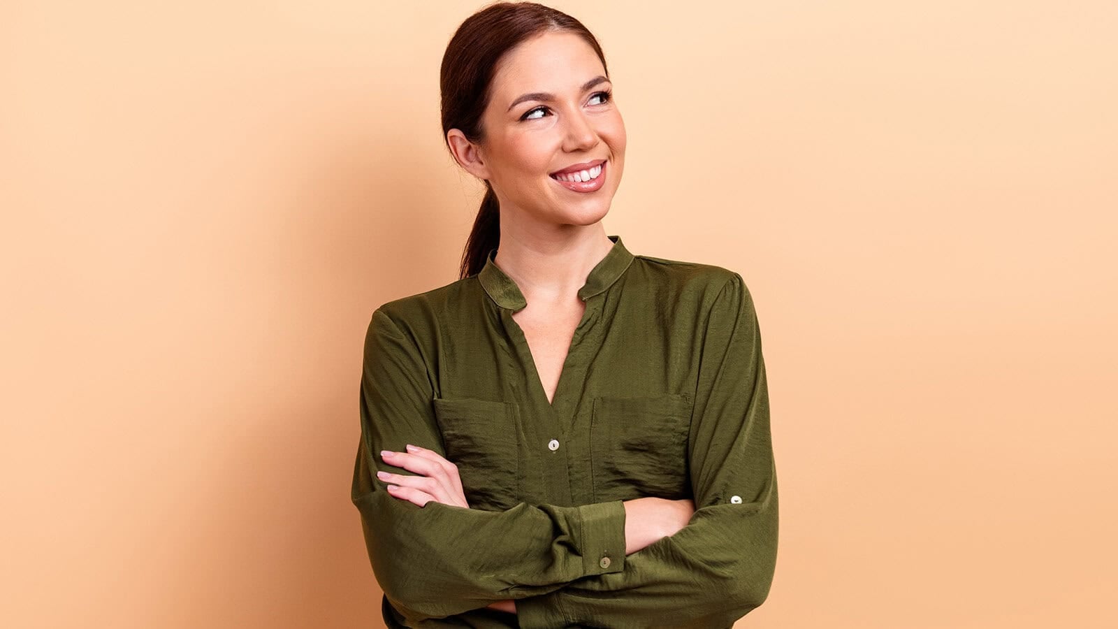 Smiling woman with brown hair in a ponytail, wearing an olive green shirt, stands with arms crossed and looks to her left against a beige background.
