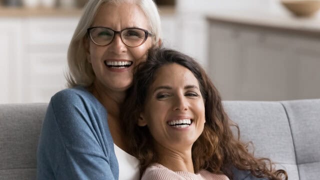 Two women, one older with gray hair and glasses, and one younger with long brown hair, sit closely together on a couch, smiling and laughing in a bright, cozy living room.