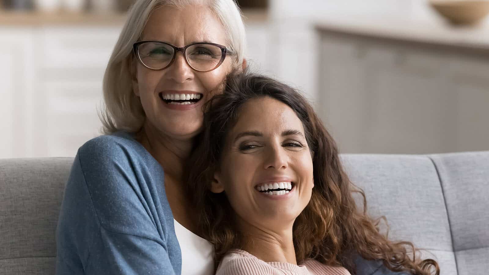Two women, one older with gray hair and glasses, and one younger with long brown hair, sit closely together on a couch, smiling and laughing in a bright, cozy living room.