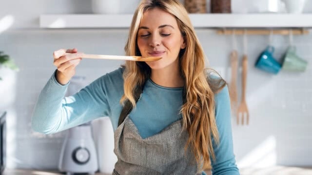 A woman wearing a gray apron tastes food from a wooden spoon in a bright kitchen, smiling with her eyes closed.