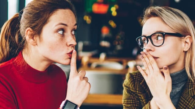 Two women sitting indoors. One woman, wearing a red sweater, holds a finger to her lips, signaling shh. The other woman, wearing glasses and a plaid jacket, looks at her with a hand partly covering her mouth.