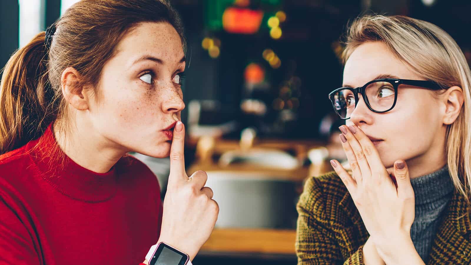 Two women sitting indoors. One woman, wearing a red sweater, holds a finger to her lips, signaling shh. The other woman, wearing glasses and a plaid jacket, looks at her with a hand partly covering her mouth.
