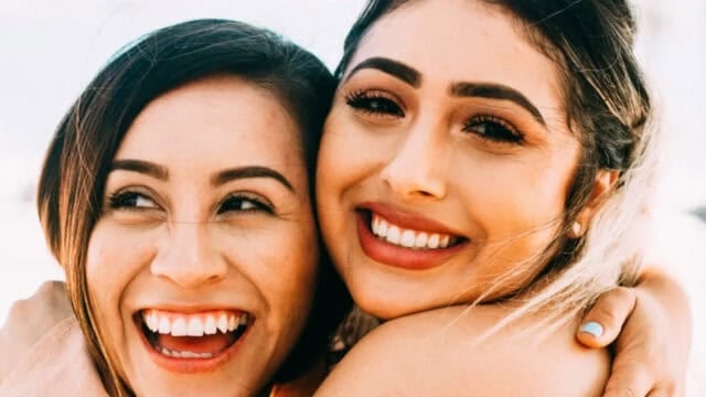 Two women smiling and hugging closely, showing their teeth and happiness, with bright natural light in the background.