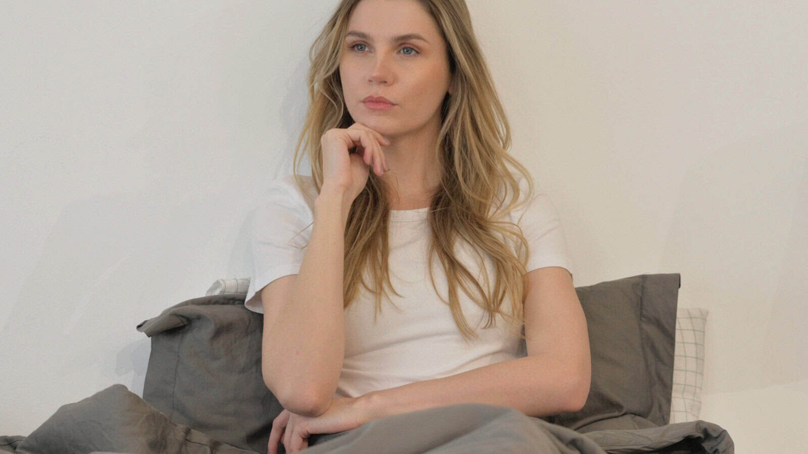 A woman with long blonde hair sits on a bed with gray bedding, wearing a white t-shirt, resting her chin on her hand and looking thoughtful.