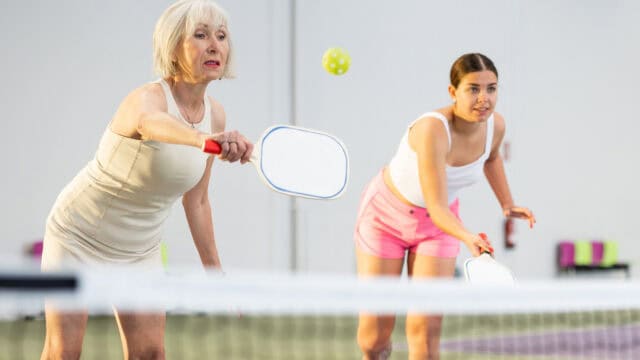 Two women playing pickleball indoors, both focused on the ball. The older woman in beige reaches to hit the ball, while the younger woman in pink shorts prepares behind her.