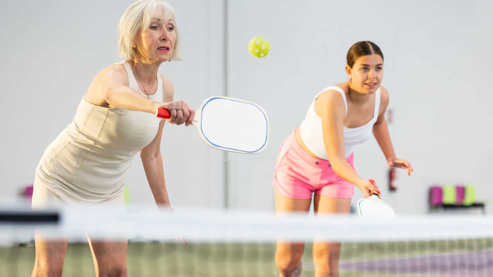 Two women playing pickleball indoors, both focused on the ball. The older woman in beige reaches to hit the ball, while the younger woman in pink shorts prepares behind her.