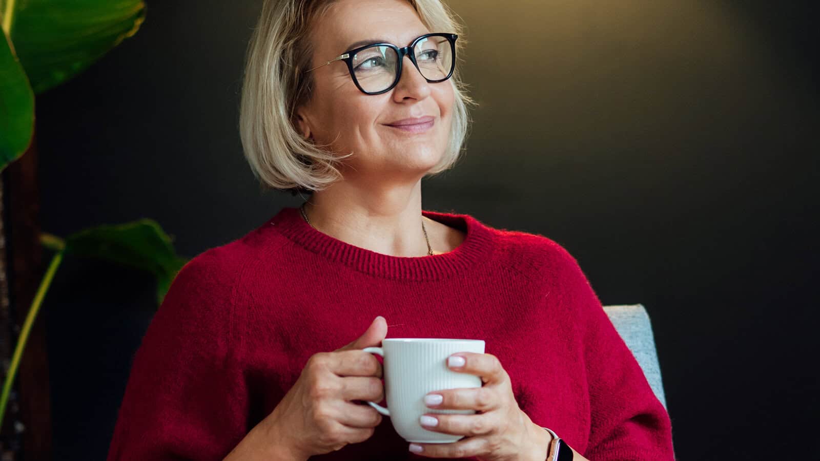 A woman with short blonde hair and glasses, wearing a red sweater, sits indoors holding a white mug and smiling thoughtfully.