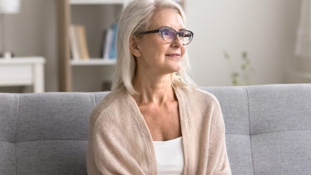 Older woman with gray hair and glasses, wearing a light sweater, sits on a gray sofa and looks thoughtfully to the side in a bright, cozy living room.