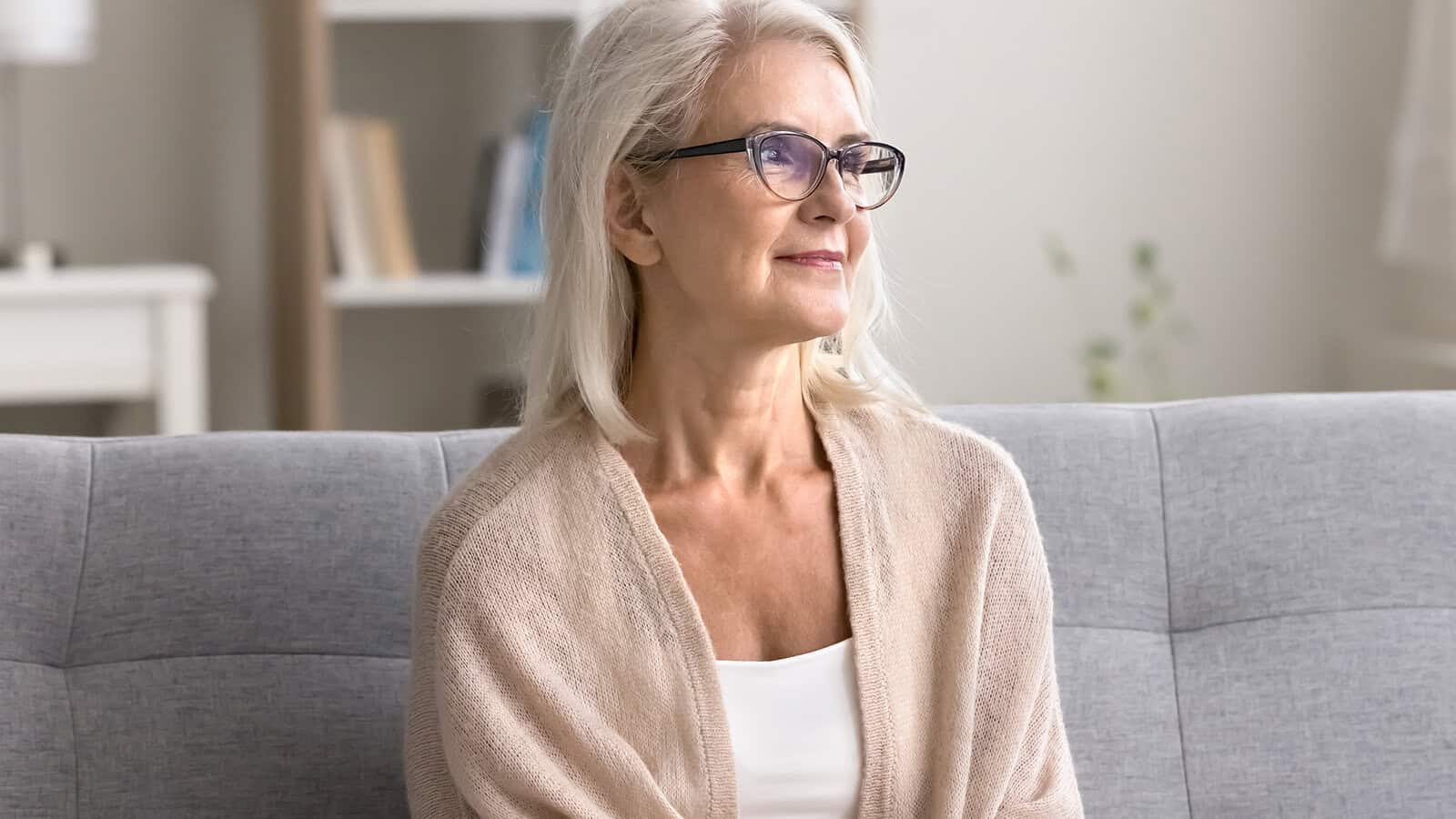 Older woman with gray hair and glasses, wearing a light sweater, sits on a gray sofa and looks thoughtfully to the side in a bright, cozy living room.