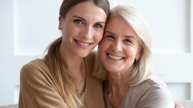 Two women, one younger and one older, are sitting close together, smiling warmly at the camera. They both have light hair and appear happy and affectionate, suggesting a close relationship, possibly mother and daughter.