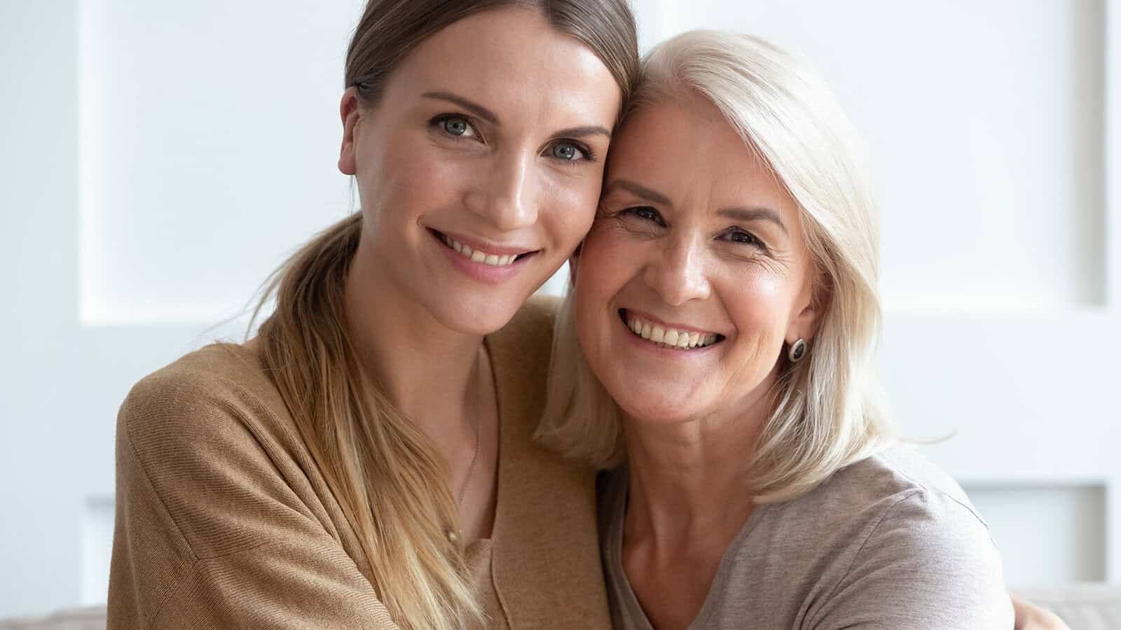 Two women, one younger and one older, are sitting close together, smiling warmly at the camera. They both have light hair and appear happy and affectionate, suggesting a close relationship, possibly mother and daughter.