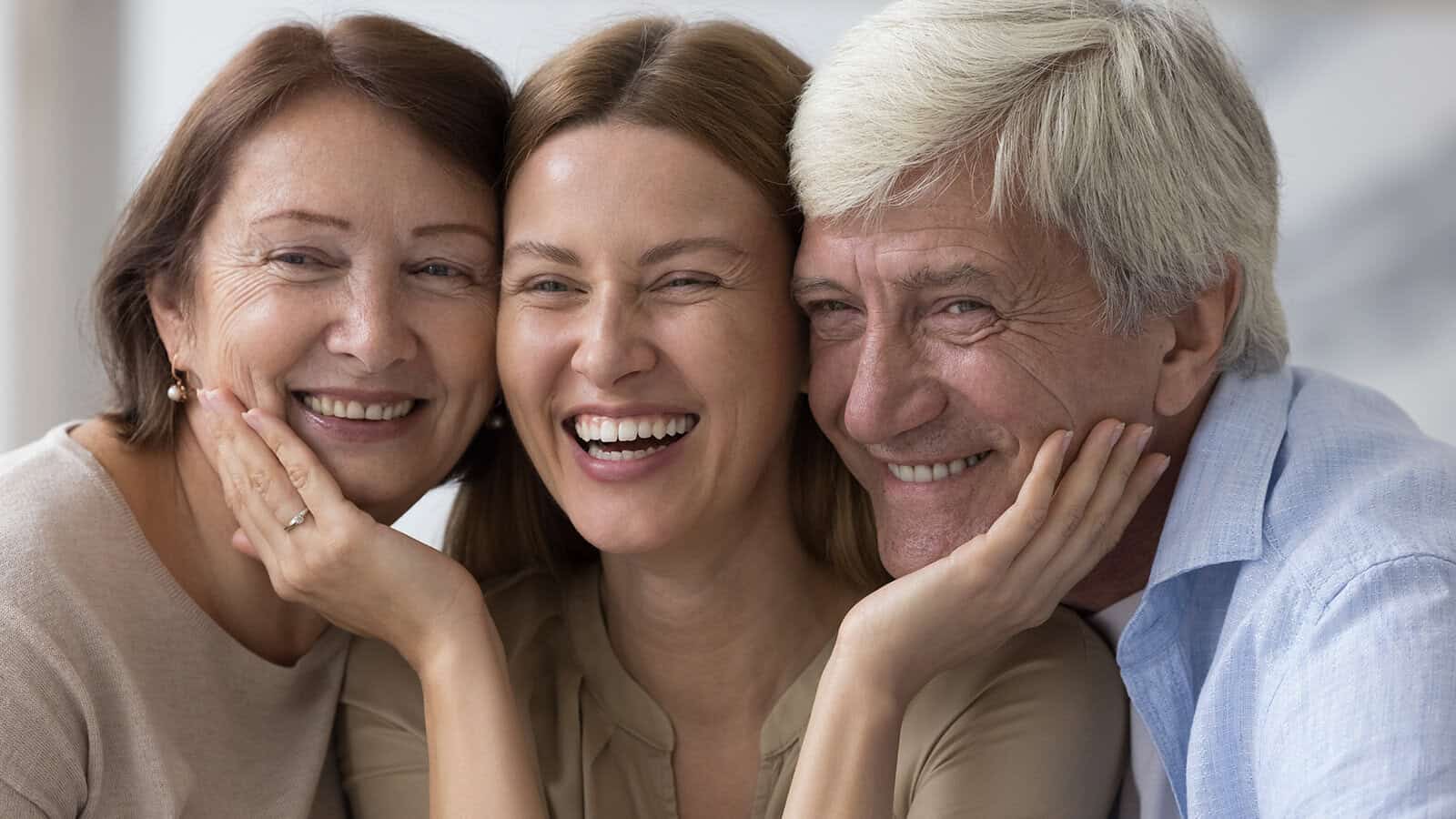 Three people smiling closely together, with a younger woman in the center and two older adults on either side, all appearing happy and affectionate.