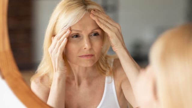 A middle-aged woman with blonde hair looks at herself in the mirror, gently touching her forehead with both hands, appearing thoughtful or concerned about her skin.