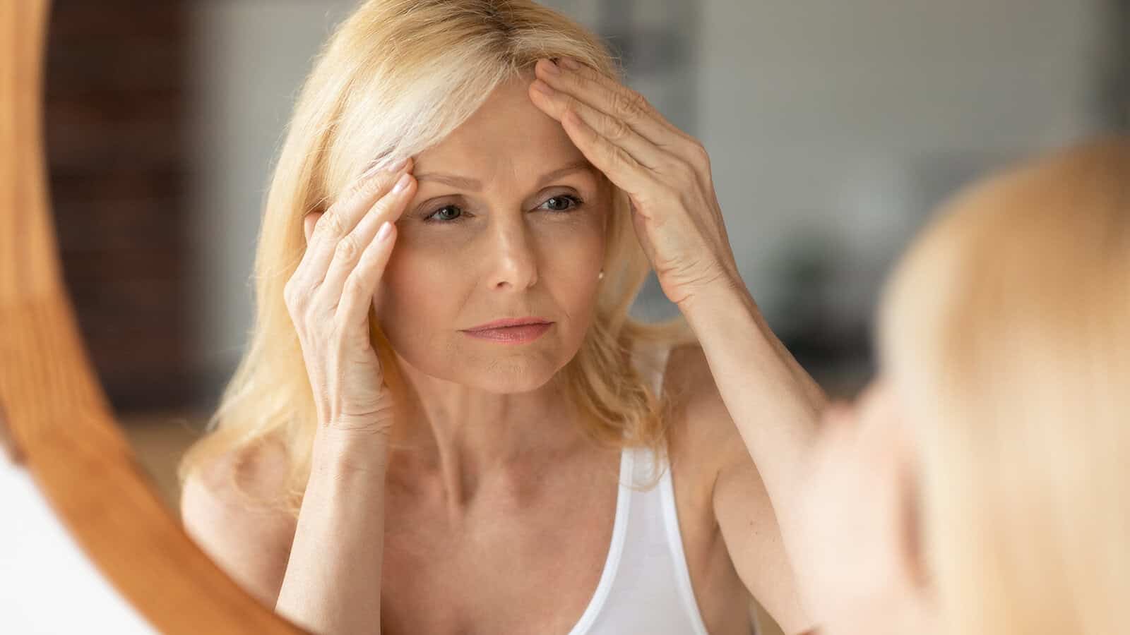 A middle-aged woman with blonde hair looks at herself in the mirror, gently touching her forehead with both hands, appearing thoughtful or concerned about her skin.