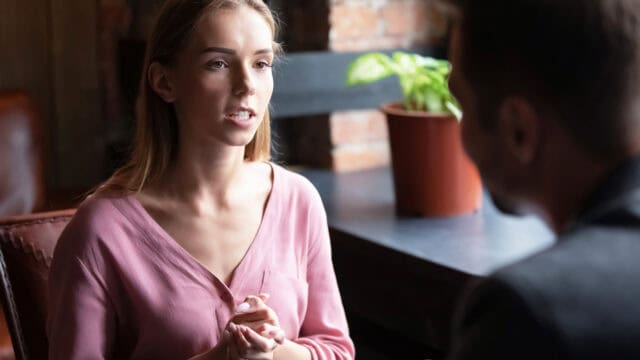 A woman in a pink top sits indoors, talking to a man whose back is to the camera. She looks engaged and is clasping her hands. There is a potted plant and brick wall in the background.