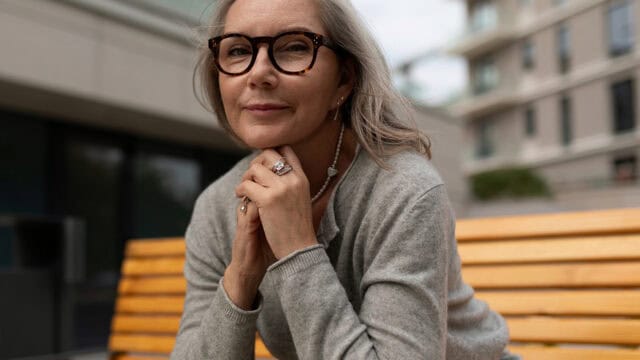 A woman with glasses and gray hair sits on a wooden bench outdoors, resting her chin on her hands and smiling softly. She wears a gray sweater, rings, and a pearl necklace. The background is blurred buildings.