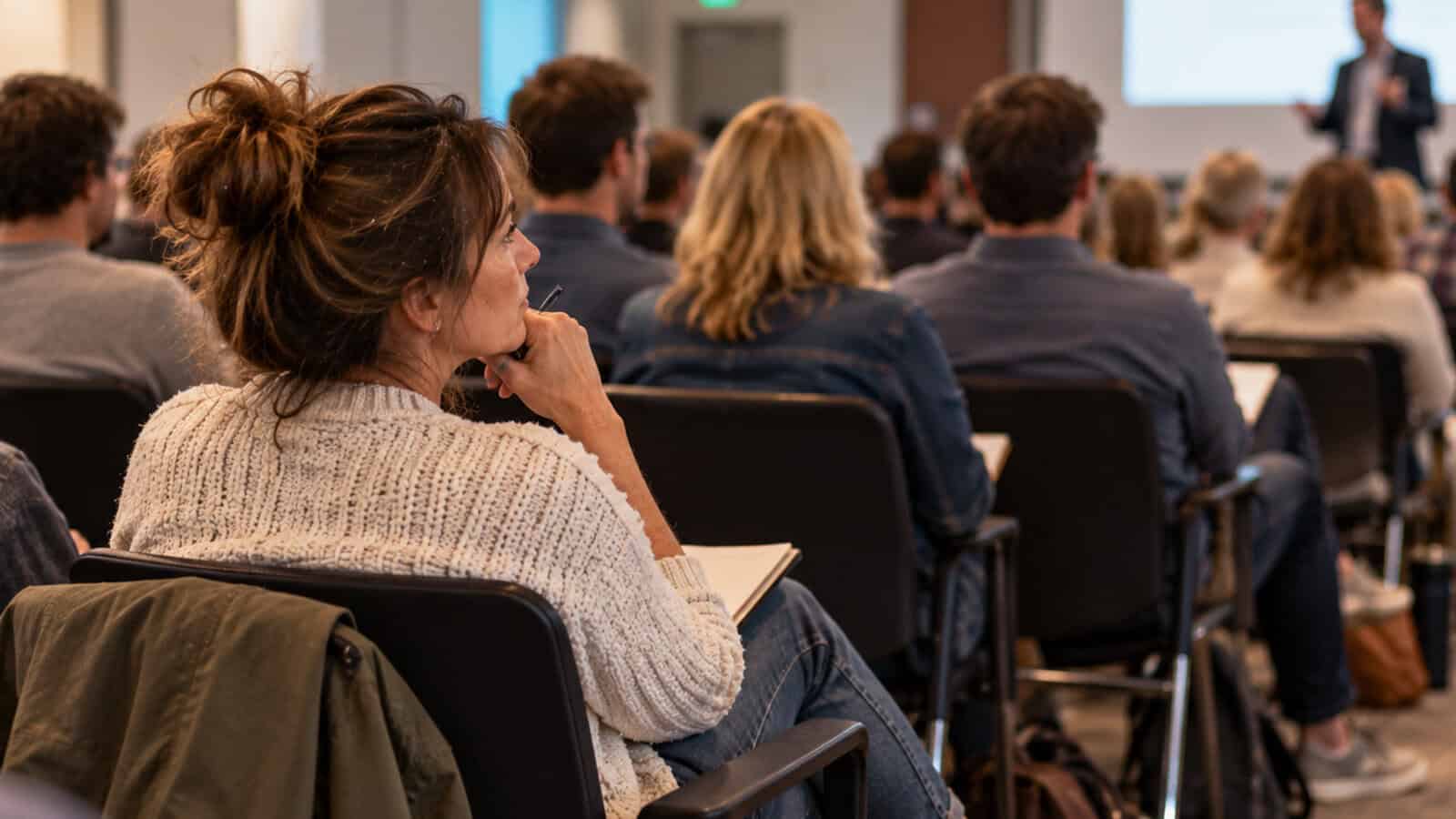A woman in a sweater sits among a group of people in a classroom or seminar, listening attentively to a speaker at the front of the room.
