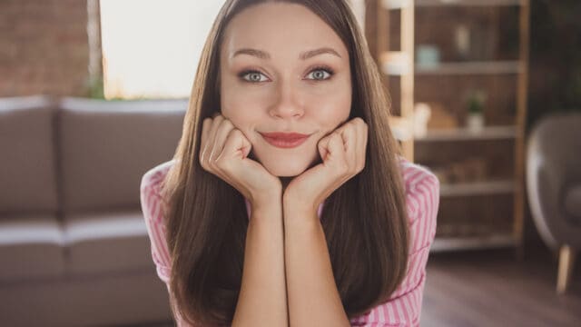 A woman with long brown hair rests her chin on her hands and smiles softly at the camera, sitting indoors with a blurred sofa and shelves in the background.