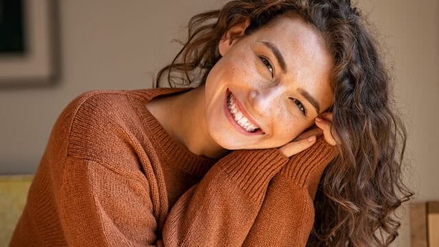 A woman with curly brown hair wearing a rust-colored sweater smiles brightly, resting her head on her hands.
