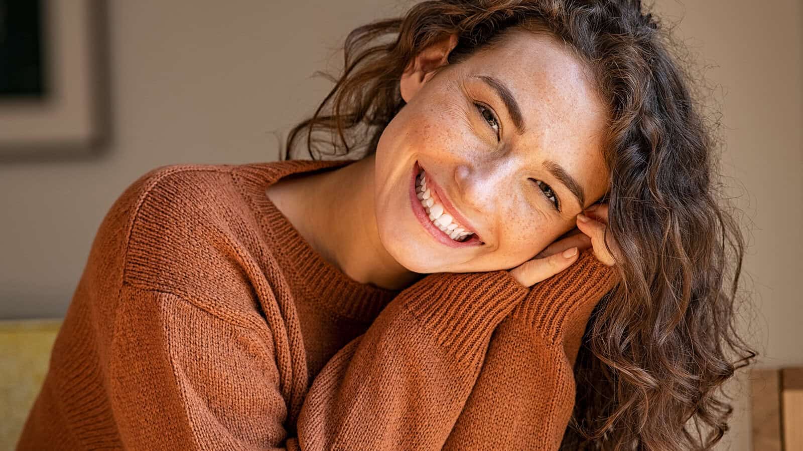 A woman with curly brown hair wearing a rust-colored sweater smiles brightly, resting her head on her hands.