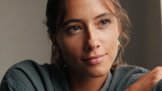 A woman with light brown hair and hazel eyes looks thoughtfully to the side, wearing a dark shirt and gold hoop earrings, with soft lighting on her face.