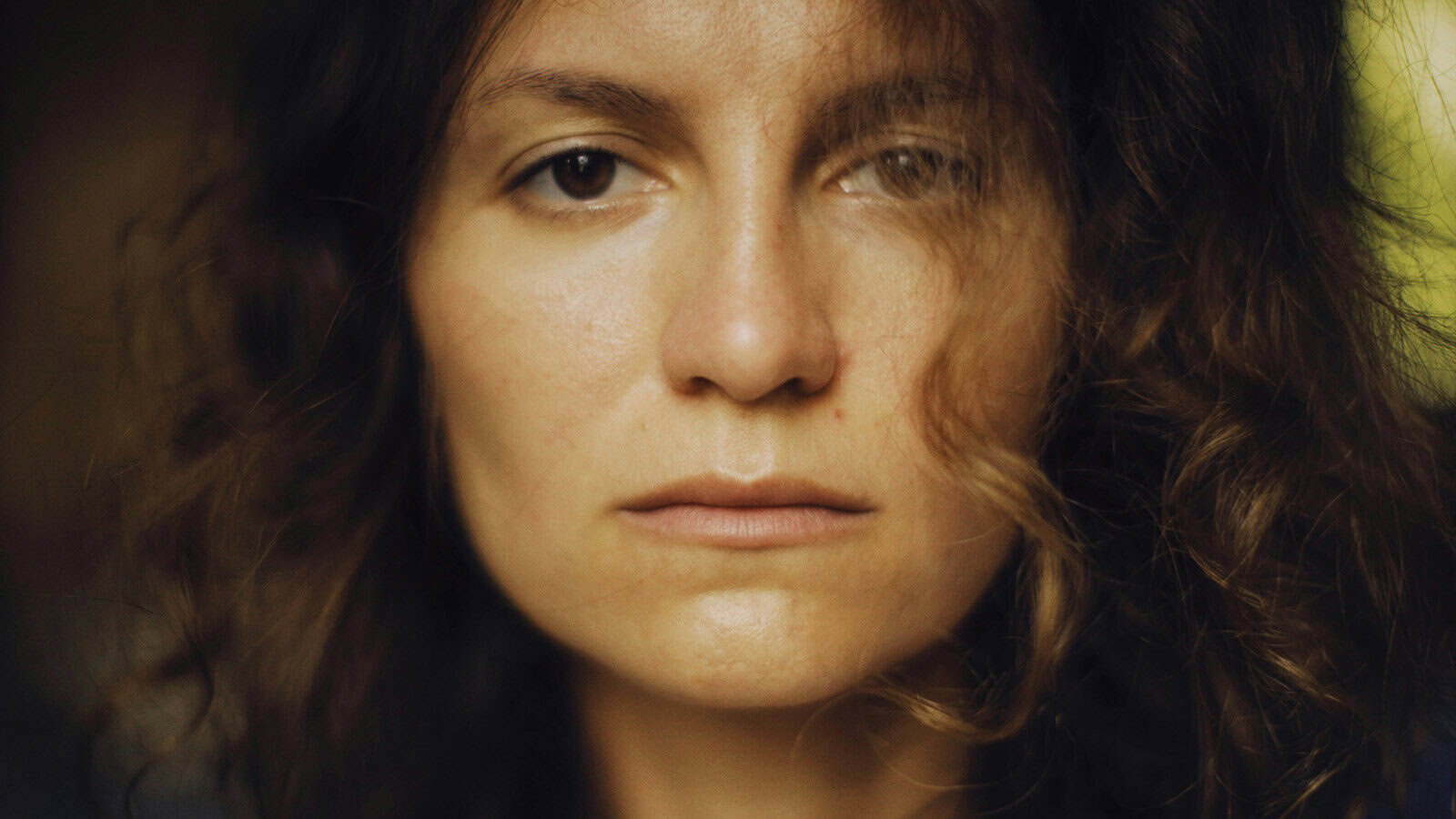 A person with wavy brown hair looks directly at the camera with a neutral expression. Their face is softly lit, and part of their hair falls across their face.