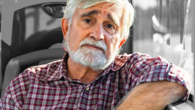 An older man with gray hair and beard, wearing a red plaid shirt, sits indoors and looks thoughtfully at the camera, resting his arm on a seat beside a window.