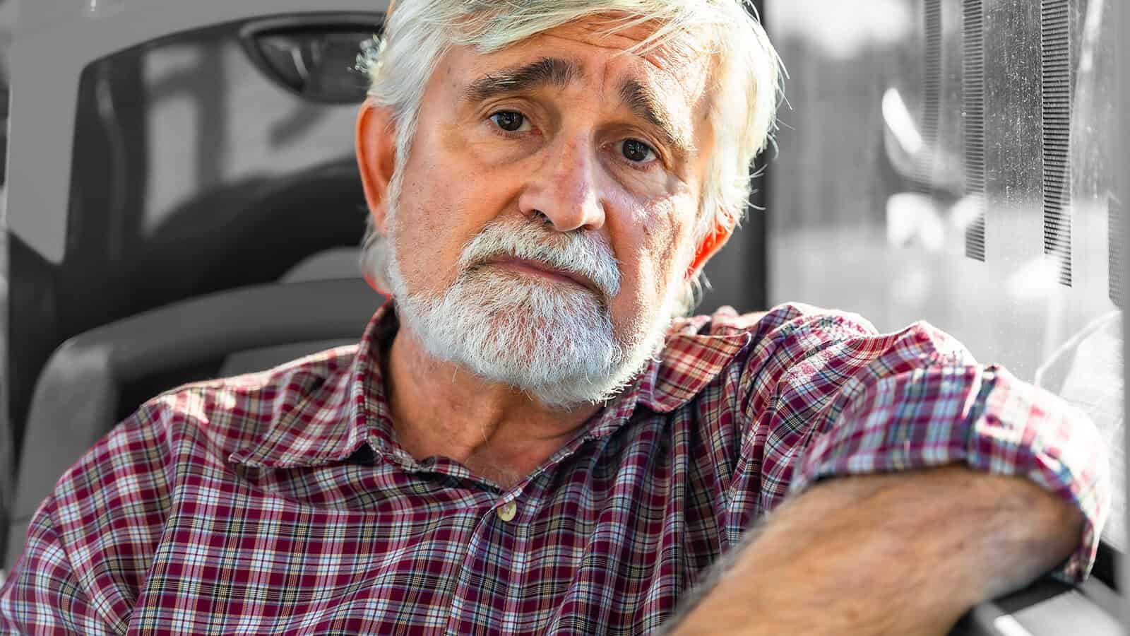 An older man with gray hair and beard, wearing a red plaid shirt, sits indoors and looks thoughtfully at the camera, resting his arm on a seat beside a window.