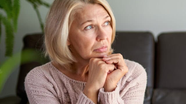 A woman with short blonde hair and a pink sweater sits indoors, resting her chin on her hands and looking thoughtfully to the side.