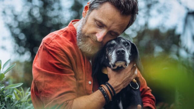 A man in an orange shirt lovingly hugs a black dog outdoors, both appearing calm and content, with greenery in the blurred background.