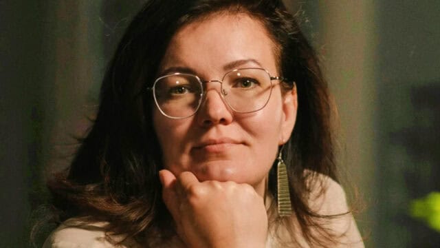 Woman with glasses and long brown hair rests her chin on her hand, looking at the camera. She wears a beige top, dangling earrings, and appears thoughtful. The background is softly blurred.